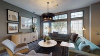 This single-floor amenity space is thoughtfully designed to serve as both a social hub and a functional work environment. The lounge area immediately draws attention with its dramatic feathered chandelier, a sculptural centerpiece that anchors the room and sets a sophisticated, design-forward tone throughout. Soft sage-green and muted gray walls complement the dark, patterned carpeting underfoot, creating a refined yet welcoming atmosphere.

The primary lounge features generously sized teal velvet sectional sofas and coordinating upholstered accent chairs arranged around a round marble-top coffee table. Large windows with plantation shutters allow ample natural light to filter in, offering views of the surrounding greenery. A light wood credenza and curated framed artwork complete the lounge's polished aesthetic.

Adjacent to the lounge, a built-in business center provides multiple computer workstations along a continuous wall-mounted desk, illuminated by individual sconce lighting and decorative framed art pieces. A dedicated conference room offers a long communal table, wire-cage pendant lighting, a whiteboard, and additional artwork, making it well-suited for meetings or focused work sessions.

A glazed door connects the space directly to a fully equipped fitness center, adding convenient functionality to this well-appointed amenity suite.