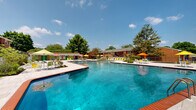 This two-floor brick residential community offers a well-rounded array of outdoor amenities set across generously landscaped grounds. The classic red brick construction is consistent throughout the multi-building complex, with each building featuring exterior stairways and private balconies that provide residents with personal outdoor space.

At the heart of the community lies a standout resort-style swimming pool with crystal-clear blue water, surrounded by a spacious tiled deck. The pool area is outfitted with vibrant green lounge chairs, colorful patio umbrellas, and neatly arranged seating — ideal for relaxation on sunny days. A decorative lattice block wall borders part of the pool deck, adding architectural character while defining the space.

Beyond the pool, a gated pathway connects to the broader grounds, where mature trees provide generous shade throughout the property. The community also features a dedicated children's playground area with rubberized flooring, as well as communal seating areas with benches positioned beneath tall evergreens. Picnic tables and outdoor grills are distributed across the grounds, encouraging outdoor gathering.

Wide concrete walkways wind through the landscaped open spaces, connecting the various buildings and amenity areas. The overall ambiance is that of a well-established, amenity-rich residential community that balances comfort, greenery, and communal living.