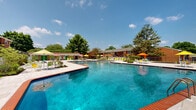 This two-floor brick residential community offers a well-rounded array of outdoor amenities set across generously landscaped grounds. The classic red brick construction is consistent throughout the multi-building complex, with each building featuring exterior stairways and private balconies that provide residents with personal outdoor space.

At the heart of the community lies a standout resort-style swimming pool with crystal-clear blue water, surrounded by a spacious tiled deck. The pool area is outfitted with vibrant green lounge chairs, colorful patio umbrellas, and neatly arranged seating — ideal for relaxation on sunny days. A decorative lattice block wall borders part of the pool deck, adding architectural character while defining the space.

Beyond the pool, a gated pathway connects to the broader grounds, where mature trees provide generous shade throughout the property. The community also features a dedicated children's playground area with rubberized flooring, as well as communal seating areas with benches positioned beneath tall evergreens. Picnic tables and outdoor grills are distributed across the grounds, encouraging outdoor gathering.

Wide concrete walkways wind through the landscaped open spaces, connecting the various buildings and amenity areas. The overall ambiance is that of a well-established, amenity-rich residential community that balances comfort, greenery, and communal living.