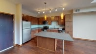Located on Excelsior Boulevard in St. Louis Park, Minnesota, this two-story residence delivers a thoughtfully designed living experience with contemporary finishes throughout. The main level showcases rich hardwood flooring and an open-concept kitchen outfitted with warm wood cabinetry, a mosaic tile backsplash, granite-style countertops, stainless steel appliances, and a central island with pendant lighting — ideal for both everyday cooking and entertaining. A track lighting system adds a modern touch overhead. The living area extends to a private balcony through floor-to-ceiling sliding glass doors, framing lush outdoor views and flooding the space with natural light. Soaring double-height ceilings amplify the sense of openness. The upper level features a carpeted bedroom area and direct views down to the living space below, creating an airy loft-like connection. A large wooden surface along the upper level overlooks the neighborhood through dual roller-shade windows. The space includes two well-appointed bathrooms: one with a sleek vanity, tile flooring, and a glass-enclosed shower, and another featuring an extended vanity, soaker tub, and tall wood cabinetry offering generous storage. Clean neutral tones and consistent wood accents throughout contribute to a cohesive, modern aesthetic.