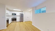 This thoughtfully designed 2-bedroom, 1-bathroom residence offers a functional and modern living experience across a single floor. The open living area is finished with light wood-style flooring and crisp white walls, illuminated by multiple windows fitted with slatted blinds that allow natural light to filter through. The kitchen is a standout feature, showcasing white shaker-style cabinetry, a classic subway tile backsplash, white quartz countertops, and a full suite of stainless steel appliances including a refrigerator, range, over-the-range microwave, and dishwasher. A double-door closet adjacent to the kitchen provides convenient additional storage. Both bedrooms are carpeted and feature neutral walls with windows that bring in natural light. The primary bedroom is generously sized and includes a ceiling fan with light fixture, while the second bedroom offers a quieter, more compact layout. The bathroom is neatly finished with large-format gray tile surrounding the tub-shower combination, brushed nickel fixtures, a white vanity, and wood-style flooring. A hallway and dedicated closet complete the layout, offering practical organization throughout this clean and move-in-ready residence.