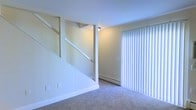 This two-story residence is thoughtfully laid out across two levels connected by a staircase visible from the main living area. The living space on the ground floor features wall-to-wall carpeting and neutral, light-toned walls that lend a clean, understated aesthetic. A large sliding glass door fitted with vertical blinds allows natural light to filter into the room while offering potential access to an outdoor area. A baseboard heating unit runs along the wall, providing an efficient heating solution. The staircase, finished in white with an open-railing design, serves as a functional architectural element within the space.

The kitchen is designed in a classic galley configuration, with cabinetry and appliances lining both sides of a central walkway. Warm-toned wood cabinetry provides ample storage above and below the dark speckled countertops. The kitchen is equipped with a stainless steel sink, dishwasher, refrigerator, and an electric range. Light hardwood-style flooring runs the length of the kitchen, and a large skylight panel in the drop ceiling floods the space with bright, even overhead illumination. Baseboard heating is also present in the kitchen, ensuring consistent comfort throughout.