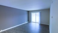 This single-floor space showcases a clean, modern aesthetic with a thoughtfully designed open living area. The room is finished with rich, dark hardwood flooring that provides a striking contrast against the soft gray walls and crisp white trim and baseboards. A large sliding glass door serves as a focal point, flooding the interior with natural light and offering a view to the outdoors, while sheer white curtains add a touch of softness to the space. The ceiling is bright white, enhancing the airy, open feel of the room. The overall layout is spacious and versatile, providing a blank canvas suitable for a variety of furniture arrangements. The neutral color palette throughout creates a calm and inviting ambiance, making the space feel both modern and welcoming. The combination of the dark flooring, muted wall tones, and generous natural light gives the space a well-balanced, contemporary character that would appeal to a wide range of preferences.