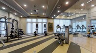 This fitness center is designed to provide a comprehensive workout environment within a single-level layout. The space is bright and welcoming, with a row of large windows dressed in sheer curtains that allow abundant natural light to fill the room. Two ceiling fans are mounted within a tray ceiling finished in a deep gray tone, complemented by recessed lighting that ensures even illumination throughout. The flooring features a bold striped pattern in alternating shades of gray and cream, adding a dynamic visual element to the space. A floor-to-ceiling mirror runs along one wall, effectively doubling the perceived size of the room and allowing users to monitor their form during exercise. Equipment includes multiple elliptical trainers and treadmills positioned near the windows, a cable weight machine, and a free weight rack along one side. Framed artwork on the walls adds a refined touch, softening the utilitarian nature of the space. The overall ambiance strikes a balance between functional and polished, creating an inviting atmosphere well-suited for a variety of fitness activities.