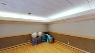 This single-level fitness and yoga studio is thoughtfully designed to support a variety of exercise and wellness activities. The space is immediately distinguished by its warm hardwood flooring, which provides an ideal surface for yoga, stretching, and floor-based workouts. The walls feature a two-tone treatment with a cork-textured lower panel accented by natural wood trim, lending the room a grounded, earthy aesthetic that promotes a calm and focused atmosphere.

Overhead, a distinctive cove lighting system runs along the perimeter of the ceiling, casting a soft, continuous glow that evenly illuminates the room without harsh shadows — ideal for a mindful exercise environment. The drop ceiling with acoustic tiles further enhances the room's suitability for group classes or individual practice by moderating sound.

A central air vent ensures proper ventilation throughout the space, maintaining comfort during active use. Neatly organized in one corner, a collection of equipment is readily accessible, including stability exercise balls, stacked foam and fabric exercise mats in multiple colors, rolled yoga mats stored in a rack, and a wicker storage basket for smaller accessories. The open floor plan provides ample room for movement, making this a well-appointed and functional space for fitness, yoga, or group wellness programming.