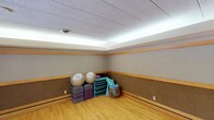This single-level fitness and yoga studio is thoughtfully designed to support a variety of exercise and wellness activities. The space is immediately distinguished by its warm hardwood flooring, which provides an ideal surface for yoga, stretching, and floor-based workouts. The walls feature a two-tone treatment with a cork-textured lower panel accented by natural wood trim, lending the room a grounded, earthy aesthetic that promotes a calm and focused atmosphere.

Overhead, a distinctive cove lighting system runs along the perimeter of the ceiling, casting a soft, continuous glow that evenly illuminates the room without harsh shadows — ideal for a mindful exercise environment. The drop ceiling with acoustic tiles further enhances the room's suitability for group classes or individual practice by moderating sound.

A central air vent ensures proper ventilation throughout the space, maintaining comfort during active use. Neatly organized in one corner, a collection of equipment is readily accessible, including stability exercise balls, stacked foam and fabric exercise mats in multiple colors, rolled yoga mats stored in a rack, and a wicker storage basket for smaller accessories. The open floor plan provides ample room for movement, making this a well-appointed and functional space for fitness, yoga, or group wellness programming.