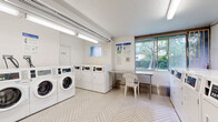 Located at 739 Birchmount Rd in Toronto's Scarborough district, this shared laundry facility comprises two laundry rooms across a single floor, offering a practical and functional space for residents. The facility is outfitted with multiple commercial-grade Coinamatic front-loading washers and top-loading dryers, arranged in neat rows along white-painted walls. The floors throughout feature a classic herringbone-patterned tile, lending a clean and orderly aesthetic to the space. Bright fluorescent lighting ensures excellent visibility throughout both rooms. One laundry room includes a stainless steel folding table paired with seating, providing a convenient area for sorting and folding laundry. A large window in this area allows natural light to filter in, offering a pleasant view of the surrounding greenery outside. Instructional signage for washer and dryer usage is clearly posted on the walls, alongside a fire extinguisher for safety compliance. The white walls and light-toned flooring create a bright, hygienic atmosphere throughout. The layout is efficient and spacious enough to accommodate multiple users simultaneously, making this a well-organized and fully functional communal laundry amenity.