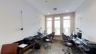 This single-floor business center is thoughtfully designed to provide a functional and professional workspace environment. The room features multiple dark-wood desks arranged to maximize usability, each equipped with computer monitors and accompanied by ergonomic high-back leather chairs, ensuring comfort during extended work sessions. A printer and additional office equipment are conveniently stationed along a dedicated side counter, catering to a range of business needs. The space is bathed in natural light streaming through a trio of large windows fitted with adjustable horizontal blinds, creating a bright yet controlled atmosphere. Recessed ceiling lighting supplements the natural light, ensuring consistent illumination throughout. The neutral beige walls and patterned low-pile carpet tiles lend the room a clean, professional aesthetic. Ceiling height adds a sense of openness to the workspace. The overall ambiance is organized and efficient, making this business center well-suited for remote work, meetings, or everyday administrative tasks.