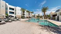 Located in Goodyear, Arizona, this outdoor amenity space is designed to deliver a resort-style experience under the Arizona sun. At the heart of the area is a generously sized swimming pool with crystal-clear water, complemented by a separate spa and a striking water feature wall finished in mosaic tile, creating a visually engaging focal point. The pool surround is finished in large-format stone pavers that extend across a broad, open deck, providing ample space for relaxation and socializing. Rows of contemporary lounge chairs and chaise lounges are arranged throughout the deck, accompanied by shade umbrellas that offer respite from the desert heat. Mature date palms are strategically placed around the perimeter, lending a tropical ambiance while framing views of the multi-story residential buildings that border the space. A covered pergola structure provides additional shaded gathering space at the far end of the deck. The area is enclosed by a wrought iron fence, ensuring privacy and security while maintaining an open, airy feel. Lush green landscaping and manicured shrubs accent the hardscape, softening the overall aesthetic. The combination of water elements, shade structures, and thoughtful landscaping makes this outdoor space a standout communal amenity.