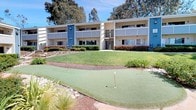 This inviting residential community is designed around a beautifully maintained central courtyard that serves as the heart of the property. The multi-building complex features a classic two-story architectural style with blue and neutral-toned siding, open-air balconies, and staircase access connecting the levels. Lush, carefully curated landscaping frames the grounds, incorporating manicured lawns, ornamental shrubs, decorative mulch beds, and native plantings that add vibrant texture throughout the outdoor space. A standout feature of the community is the dedicated putting green, offering a fun and unique recreational amenity for residents. A pergola-covered patio provides a shaded outdoor gathering area complete with wicker dining furniture, ideal for al fresco meals or casual socializing. Nearby, a stainless steel gas grill station adds to the outdoor entertaining options. Meandering pathways connect the various areas of the courtyard, encouraging a leisurely stroll through the grounds. Towering mature trees line the perimeter, providing natural shade and a sense of privacy. The overall ambiance is bright, airy, and welcoming, with abundant natural sunlight enhancing the open, park-like setting of this thoughtfully designed community.