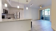 This single-floor residence offers a thoughtfully designed open-concept layout that seamlessly connects the kitchen and living areas. The kitchen serves as the centerpiece of the space, showcasing a generous granite-topped island with a built-in sink, ideal for both meal preparation and casual dining. Crisp white cabinetry lines the walls, complemented by stainless steel appliances, including a full-sized refrigerator, creating a clean and contemporary aesthetic. Pendant lighting hangs above the island, adding a warm, focused glow, while track lighting along the ceiling provides versatile illumination throughout. Light-toned tile flooring runs consistently across the open-plan area, enhancing the sense of continuity and spaciousness. The living area extends naturally from the kitchen, bathed in natural light streaming in through a full-width sliding glass door that opens to a private balcony, offering a seamless indoor-outdoor connection. The high ceilings amplify the airy feel of the space, while the neutral color palette throughout creates a bright, welcoming ambiance. Overall, this residence combines modern finishes with a functional, open layout, making it an inviting and versatile living environment.