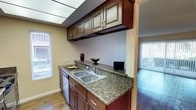 This single-floor residence offers a thoughtfully designed living space that blends style and functionality. The kitchen serves as a standout feature, showcasing rich, dark-stained cabinetry with raised-panel detailing that extends both above and below the countertops. The granite countertops provide ample prep space, complemented by a double-basin stainless steel sink and built-in dishwasher. A skylight-style ceiling fixture brightens the kitchen workspace, while a window with blinds allows for natural light. The warm, wood-toned cabinetry is accented by brushed metal hardware, adding a refined touch to the overall aesthetic. Hardwood flooring flows seamlessly from the kitchen into the adjacent living area, creating a cohesive and inviting atmosphere throughout the space. Vertical blinds on a sliding glass door in the living area suggest access to an outdoor area, allowing for additional natural light. The neutral wall tones provide a versatile backdrop, complementing the warm woodwork and stone surfaces throughout. The overall design balances practicality with comfort, making this a well-suited space for everyday living.