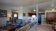 This single-floor space features an open-concept design that seamlessly connects the living area and kitchen, creating a cohesive and functional layout. The living area is furnished with a plush sofa and a patterned armchair, arranged to encourage comfortable seating and conversation. A small side table with a lamp adds a warm glow, complementing the neutral carpet flooring and light-colored walls throughout.

Vaulted ceilings rise above the kitchen area, lending an airy sense of openness. The kitchen is equipped with medium-toned wooden cabinetry, a white refrigerator, and a countertop with bar-style seating featuring wrought-iron stools — ideal for casual dining. A coffee maker and small appliances are neatly arranged along the counter.

A structural column subtly defines the transition between the living and kitchen spaces without disrupting the open feel. A framed piece of wall art adds a decorative touch to the living area. A doorway leads to an additional room, suggesting further private accommodations. The overall ambiance is warm, understated, and practical, making the space feel inviting and well-suited for everyday living.