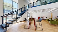 This two-story commercial space combines contemporary design with practical functionality across a well-organized layout. The ground floor makes an immediate impression with its soaring double-height ceilings and a dramatic stacked-stone accent wall that stretches upward, anchored by a striking multi-arm sputnik chandelier overhead. A reception desk sits at the entry alongside large windows that frame outdoor greenery, flooding the space with natural light. Light-toned hardwood flooring runs throughout the open area, which accommodates a mix of seating arrangements, including counter-height tables with modern chairs and a lounge zone furnished with mid-century-inspired olive green armchairs. A kitchenette with wood cabinetry and a sink provides convenient amenities nearby.

A bold open-riser staircase with blue-patterned carpet treads and sleek black metal railings connects the ground floor to an upper mezzanine level, visible from below through glass balustrades. Exposed white-painted structural beams traverse the ceilings on both levels, contributing to the industrial-modern aesthetic. Dedicated workstation areas feature long white desks set against a vivid cobalt blue accent wall, with patterned carpet tiles and glass partition walls defining semi-private office zones. Throughout, recessed and linear lighting ensures consistent illumination across all work and collaborative areas.