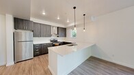 Located at 909 Eastside St SE in Olympia, Washington, this single-story, one-bedroom, one-bathroom unit offers a thoughtfully designed open-concept layout. The combined kitchen and living area forms the heart of the space, showcasing a modern aesthetic defined by dark wood-grain cabinetry, white quartz countertops, and stainless steel appliances including a refrigerator and range with a wall-mounted hood vent. A peninsula with an integrated sink and pendant lighting serves as a functional divider between the kitchen and living areas, adding both style and practicality. Light wood-look flooring runs throughout the main living spaces, contributing to a clean, contemporary feel. Large windows in the living area flood the space with natural light and offer views of the surrounding neighborhood greenery. A stacked washer and dryer unit is conveniently built into a dedicated alcove. The bedroom features soft carpeting, a generous closet, and a window overlooking outdoor greenery. The bathroom includes wood-tone cabinetry with a white countertop vanity and wood-look tile flooring. The unit also features a private entry door with a sidelight window.