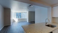 This single-floor space showcases a clean, contemporary design with an open-concept layout that seamlessly connects the kitchen and living areas. A granite countertop anchors the kitchen space, featuring a speckled beige-and-gold finish that adds a touch of elegance to the neutral palette throughout. The living area is finished with dark hardwood-style flooring that contrasts beautifully against the bright white walls, lending a modern and polished aesthetic.

Natural light floods the space through a large picture window that frames an impressive city skyline view, with surrounding high-rise buildings visible in the background — a defining feature of this urban setting. The abundance of natural light enhances the open and airy feel of the interior. Track lighting mounted on the ceiling provides additional illumination, offering both functional and accent lighting options.

The layout includes a defined corridor or secondary room visible beyond the main living space, suggesting a thoughtfully zoned floor plan. Overall, the space carries a minimalist, move-in-ready ambiance suited for urban living, characterized by neutral tones, quality finishes, and a strong connection to the surrounding cityscape.