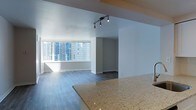 This single-floor space showcases a clean, contemporary design with an open-concept layout that seamlessly connects the kitchen and living areas. A granite countertop anchors the kitchen space, featuring a speckled beige-and-gold finish that adds a touch of elegance to the neutral palette throughout. The living area is finished with dark hardwood-style flooring that contrasts beautifully against the bright white walls, lending a modern and polished aesthetic.

Natural light floods the space through a large picture window that frames an impressive city skyline view, with surrounding high-rise buildings visible in the background — a defining feature of this urban setting. The abundance of natural light enhances the open and airy feel of the interior. Track lighting mounted on the ceiling provides additional illumination, offering both functional and accent lighting options.

The layout includes a defined corridor or secondary room visible beyond the main living space, suggesting a thoughtfully zoned floor plan. Overall, the space carries a minimalist, move-in-ready ambiance suited for urban living, characterized by neutral tones, quality finishes, and a strong connection to the surrounding cityscape.