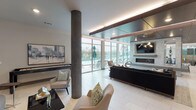 This contemporary social lounge is designed to accommodate both relaxed gatherings and vibrant social events across a single, thoughtfully organized floor. The open-plan layout flows seamlessly from a fully equipped kitchen to several distinct seating areas, all unified by large-format porcelain tile flooring and a clean, modern aesthetic.

The kitchen area is anchored by an expansive white quartz countertop island with an undermount sink, complemented by rich wood cabinetry and stainless steel appliances. Elegant pendant lighting hangs above the island, while floor-to-ceiling glass walls flood the space with natural light and provide direct views and access to an outdoor terrace with patio seating.

The main lounge area features a striking stone-clad fireplace wall with a mounted flat-screen television, set beneath a dramatic coffered ceiling finished in dark wood paneling with integrated ambient lighting. Stylish seating arrangements — including plush sofas, accent chairs, and sculptural coffee tables — are positioned throughout, catering to both intimate conversations and larger group gatherings.

A dedicated casual nook with curved built-in bench seating, teal accent walls, and curated artwork adds a cozy, distinctive character to the space. Decorative textured wall panels, statement light fixtures, and carefully selected art pieces are found throughout, contributing to a refined yet welcoming atmosphere that encourages community and connection.