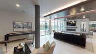 This contemporary social lounge is designed to accommodate both relaxed gatherings and vibrant social events across a single, thoughtfully organized floor. The open-plan layout flows seamlessly from a fully equipped kitchen to several distinct seating areas, all unified by large-format porcelain tile flooring and a clean, modern aesthetic.

The kitchen area is anchored by an expansive white quartz countertop island with an undermount sink, complemented by rich wood cabinetry and stainless steel appliances. Elegant pendant lighting hangs above the island, while floor-to-ceiling glass walls flood the space with natural light and provide direct views and access to an outdoor terrace with patio seating.

The main lounge area features a striking stone-clad fireplace wall with a mounted flat-screen television, set beneath a dramatic coffered ceiling finished in dark wood paneling with integrated ambient lighting. Stylish seating arrangements — including plush sofas, accent chairs, and sculptural coffee tables — are positioned throughout, catering to both intimate conversations and larger group gatherings.

A dedicated casual nook with curved built-in bench seating, teal accent walls, and curated artwork adds a cozy, distinctive character to the space. Decorative textured wall panels, statement light fixtures, and carefully selected art pieces are found throughout, contributing to a refined yet welcoming atmosphere that encourages community and connection.