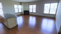 This single-floor residence offers an open and airy living space defined by rich, warm hardwood flooring that reflects natural light throughout the room. The main living area is anchored by a sleek, dark-framed fireplace set against a neutral-toned wall, providing both a functional heating element and a refined focal point. Multiple windows fitted with horizontal blinds line the walls, allowing generous amounts of natural light to fill the space while offering views of the surrounding exterior. The neutral gray and beige wall tones complement the warm flooring, creating a balanced and inviting atmosphere. A partial kitchen counter or bar-height divider is visible at the edge of the living space, featuring a dark countertop surface and light cabinetry, suggesting a semi-open kitchen layout that connects seamlessly to the main living area. The open floor plan provides a versatile and spacious feel, well-suited for various furniture arrangements. The overall ambiance is clean, modern, and move-in ready, with a cohesive design that emphasizes natural materials and understated elegance.