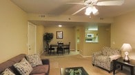 This single-floor residence is designed with an open-concept layout that seamlessly connects the living, dining, and kitchen areas into one cohesive space. The living area is comfortably appointed, featuring a plush sofa and accent chair with patterned upholstery, centered around a dark coffee table. A ceiling fan with integrated lighting fixture overhead provides both air circulation and ambient illumination throughout the space.

The dining area sits adjacent to the living room and accommodates a dark-toned dining table with matching chairs, making it well-suited for everyday meals or casual gatherings. A framed piece of wall art and a decorative indoor plant add warmth and visual interest to the shared space.

The kitchen is accessible via a pass-through opening, offering partial visibility from the main living area. It features wooden upper cabinetry, a microwave, and a range, with light-toned countertops and a functional layout. The flooring transitions from tile in the kitchen and entry areas to carpet in the living and dining zones. Warm yellow-toned walls throughout contribute to the overall inviting and comfortable ambiance of the space.