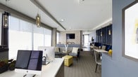 This well-appointed single-floor amenity space combines functionality and contemporary design to create an inviting, multi-use environment. The open-plan layout seamlessly integrates a business center, a relaxed lounge area, and a café-style dining nook, all unified by a cohesive modern aesthetic.

The business center area features sleek white desks equipped with desktop computers, illuminated by large windows fitted with roller shades that allow for adjustable natural light. Elegant geometric pendant light fixtures add a refined touch overhead.

Adjacent to the workspace, a comfortable lounge area centers around a tiled fireplace wall with a mounted flat-screen television above. The seating arrangement includes upholstered chairs and a sofa in neutral tones, accented by a bold yellow ottoman and a glass-topped coffee table, creating a warm and welcoming atmosphere.

Toward the rear of the space, a bar-height dining area with striped chairs and dark navy slatted wall paneling provides an intimate setting for casual gatherings. The consistent use of neutral grays, blues, and yellow accents throughout the space ties the zones together cohesively. Carpeted flooring, recessed lighting, and structural columns contribute to the space's polished, professional character.