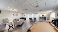Located at 10 Lucas Ln in Voorhees, NJ, this single-floor community clubhouse offers a thoughtfully designed, open-plan layout ideal for socializing and recreation. The main area is anchored by durable hardwood-style flooring and a suspended drop-tile ceiling with recessed lighting throughout, creating a bright and welcoming atmosphere. A standout fireplace with a classic white mantel serves as a focal point in the lounge area, which is complemented by a wall-mounted flat-screen TV and comfortable seating arrangements including sofas and upholstered armchairs. A dedicated billiards area features a full-size pool table, adding a recreational dimension to the space. Multiple dining tables with black chairs provide ample seating for group gatherings, while a cozy booth-style nook along one wall offers a more intimate setting. The fully equipped kitchen includes stainless steel appliances — a range, microwave, and refrigerator — white cabinetry, a classic subway tile backsplash, and light countertops. Windows along the perimeter allow natural light to fill the space. Two bathrooms and additional support rooms round out the floorplan, making this clubhouse a comprehensive and versatile community amenity.
