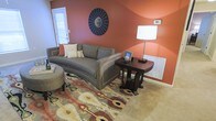 This single-floor space makes a strong first impression with its bold design choices and warm, welcoming ambiance. The living area is anchored by a striking terracotta-orange accent wall that adds depth and warmth to the room, complemented by soft neutral tones on the remaining walls. Plush wall-to-wall carpeting runs throughout, adding comfort underfoot and contributing to the cozy feel of the space. A curved gray sofa serves as the centerpiece of the living area, paired with a richly stained dark wood side table topped with a classic white lamp, creating an elegant yet relaxed seating arrangement. A vibrant multicolored area rug with an ikat-inspired pattern introduces layers of color and texture, tying the room's earthy palette together beautifully. A round decorative mirror with a starburst design adorns the accent wall, adding a modern artistic touch. Natural light filters in through a door with a window panel, brightening the space without overwhelming its warm color scheme. A wall-mounted heating unit ensures year-round comfort. The overall ambiance is one of curated coziness — a thoughtfully styled space that balances bold color with comfortable, functional furnishings.