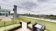 This rooftop terrace offers an impressive outdoor living experience elevated above the surrounding urban landscape. The terrace is defined by a striking living green roof system, where low-growing sedum and groundcover plants extend across a substantial portion of the surface, providing both aesthetic appeal and environmental benefits. A tiled patio area serves as the primary gathering space, furnished with dark wicker outdoor sofas and chairs fitted with vivid green cushions, arranged to maximize enjoyment of the views.

Tall, industrial-style steel columns rise from the terrace deck, fitted with wall-mounted speakers to provide ambient sound throughout the space. The perimeter is secured by a sleek glass and metal railing system, ensuring unobstructed sightlines across the cityscape. Panoramic views of a recognizable urban skyline stretch across the horizon, framed by a canopy of mature trees at lower elevations.

Access to the rooftop is provided through a glass-enclosed stairwell structure situated centrally on the terrace, featuring double glass doors and a clean, modern design. The overall ambiance is open and airy, combining natural greenery with functional outdoor furnishings to create a versatile communal rooftop retreat.