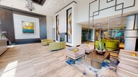 The lobby at 2827 Telek Pl in Alexandria, Virginia is a generously scaled, single-floor space that makes a striking first impression through its contemporary architectural design. Floor-to-ceiling glass at the main entrance floods the interior with natural light, highlighting the large-format travertine-style tile flooring that flows throughout. The double-height ceiling in the central atrium is punctuated by a coffered tray detail and an eye-catching sculptural chandelier, reinforcing the sense of grandeur.

The main seating area is thoughtfully curated with chartreuse accent chairs, striped upholstered armchairs, and a vibrant area rug, arranged around a sleek fireplace flanked by large-scale sculptural wall art in gold. An abstract painting adds a bold focal point to one accent wall. The concierge desk, framed by a dramatic stacked stone and backlit panel feature wall, anchors the reception area with a polished, professional aesthetic.

Wide, well-lit corridors extend from the lobby, their walls accented with geometric paneling and art niches, leading residents to elevator bays illuminated by distinctive circular pendant light fixtures. A dedicated package room with bank-style lockers and a convenient pet amenity station round out the practical offerings. Together, these spaces create a lobby that balances visual sophistication with everyday functionality.