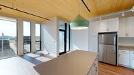 This thoughtfully designed single-floor space showcases a seamless blend of modern aesthetics and functional living. A standout architectural feature is the warm, natural wood-paneled ceiling that runs throughout, adding texture and a welcoming character to the interior while complementing the clean white walls and light hardwood flooring found across the main living areas.

The kitchen is a highlight of the space, outfitted with a full suite of stainless steel appliances including a refrigerator, electric range, built-in microwave, and dishwasher. Crisp white shaker-style cabinetry lines the walls, providing generous storage, while neutral-toned countertops offer ample workspace. A central island with a pendant light in a distinctive mint-green finish serves as both a functional prep area and a visual focal point. Track lighting along the ceiling adds both practicality and a contemporary industrial touch.

The living area is bathed in natural light, courtesy of dramatic floor-to-ceiling black-framed windows that frame views of the surrounding neighborhood and allow sunlight to pour in throughout the day. A sliding glass door provides access to an outdoor area, further connecting the interior to the outside.

The bathroom maintains a clean, minimalist design with neutral tones, featuring a walk-in shower area and straightforward, functional fixtures. Overall, the space offers a bright, airy, and modern environment.