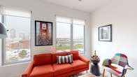 Located at 350 Mount Vernon Ave in Columbus, Ohio, this contemporary single-floor residence offers a thoughtfully designed layout with a clean, modern aesthetic throughout. The open-concept living and dining area is bright and airy, anchored by large windows that frame sweeping elevated views of the Columbus skyline and surrounding landscape, flooding the space with natural light. The living area provides a comfortable gathering space, while the adjacent dining zone offers room for a full dining set beneath a ceiling fan, creating a seamless flow between the two areas. The kitchen features a striking industrial design with stainless steel appliances, a stainless steel countertop island with bar seating, white cabinetry, subway tile backsplash, and exposed ductwork overhead, complemented by Edison bulb pendant lighting. The bedroom is generously sized with plush carpeting and large windows offering impressive city views, accessed via a sleek sliding barn door that connects to a well-appointed bathroom with clean white tilework and modern vanity. In-unit laundry with a full-size washer and dryer provides added convenience. Light hardwood-style flooring in the kitchen and living areas, combined with neutral white walls throughout, gives the entire space a bright, cohesive, and contemporary character.