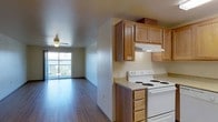 This single-floor space combines a functional kitchen and an open living area in a practical, well-organized layout. The kitchen is equipped with warm honey-toned oak cabinetry offering ample storage, a white electric coil range, light-toned countertops, and an under-cabinet range hood. The cabinetry extends to the upper walls, maximizing vertical storage space.

The kitchen flows directly into the living area, which features rich, dark hardwood-style flooring that extends throughout, creating a cohesive and visually grounded atmosphere. A ceiling fan is centrally positioned in the living area, promoting air circulation and providing overhead lighting.

A sliding glass door at the far end of the living area opens to a balcony, allowing natural light to pour into the space and offering an outdoor retreat. The textured walls and neutral color palette throughout contribute to a clean, understated aesthetic. Overall, the space is designed for comfortable, efficient everyday living, with an open feel enhanced by the connected layout and the bright outlook toward the exterior.
