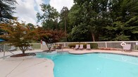 This community amenity space offers two distinct areas designed for convenience and relaxation. The outdoor pool area is a standout feature, showcasing a generously sized, freeform pool with crystal-clear blue water. The pool deck is enclosed by a white perimeter fence and is beautifully framed by mature trees and ornamental landscaping, including a striking Japanese maple. Lounge chairs are positioned poolside, providing a comfortable spot to unwind, while colorful flower beds add a welcoming, resort-like atmosphere.

The interior laundry facility is equally well-maintained and thoughtfully designed. The waiting area features a classic black-and-white checkerboard tile floor, neutral gray walls with crisp white crown molding and wainscoting, and recessed lighting. Transparent acrylic chairs offer seating while residents wait. A reading nook with a magazine rack provides added convenience. The adjacent laundry room houses multiple stacked washer and dryer units, all neatly arranged and easily accessible. Decorative artwork, including a vintage-style laundry sign and a classic French advertising poster, lend the space a charming, curated character. Natural light filters in through a window, keeping the space bright and inviting.