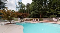 This community amenity space offers two distinct areas designed for convenience and relaxation. The outdoor pool area is a standout feature, showcasing a generously sized, freeform pool with crystal-clear blue water. The pool deck is enclosed by a white perimeter fence and is beautifully framed by mature trees and ornamental landscaping, including a striking Japanese maple. Lounge chairs are positioned poolside, providing a comfortable spot to unwind, while colorful flower beds add a welcoming, resort-like atmosphere.

The interior laundry facility is equally well-maintained and thoughtfully designed. The waiting area features a classic black-and-white checkerboard tile floor, neutral gray walls with crisp white crown molding and wainscoting, and recessed lighting. Transparent acrylic chairs offer seating while residents wait. A reading nook with a magazine rack provides added convenience. The adjacent laundry room houses multiple stacked washer and dryer units, all neatly arranged and easily accessible. Decorative artwork, including a vintage-style laundry sign and a classic French advertising poster, lend the space a charming, curated character. Natural light filters in through a window, keeping the space bright and inviting.