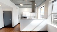 Located at 519 Market St in the heart of downtown Shreveport, Louisiana, this single-floor urban unit blends industrial character with modern finishes throughout. The open-concept kitchen serves as a focal point, featuring a large white peninsula with pendant lighting suspended from exposed concrete ceilings above. White upper cabinetry with sleek hardware pairs with dark lower cabinets accented by gold-toned pulls, complemented by white countertops, a stainless steel refrigerator, built-in dishwasher, and microwave. A statement stainless steel range hood crowns the cooking area, adding an industrial edge. Rich hardwood-style flooring runs throughout, grounding the clean white walls and raw ceiling aesthetic. The living and sleeping areas benefit from oversized windows framing sweeping views of the downtown Shreveport skyline, filling the space with abundant natural light. The bathroom is clean and functional, featuring a full tub-shower combination, wood-look flooring, and crisp white finishes with gold hardware accents. The overall ambiance is contemporary and urban, balancing raw architectural elements with polished modern touches suited to city living.