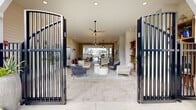 Located at 2150 NW 21st St in Ocala, Florida, this single-story community clubhouse for Aurora at Ocala presents a refined coastal aesthetic that blends indoor comfort with open-air living. The entrance is framed by striking dark metal gate doors that open to a spacious, light-filled interior finished with neutral tones and horizontal shiplap walls throughout. Stone-tile flooring extends seamlessly from the covered outdoor areas into the interior, creating a cohesive flow across the space. The main lounge area features stylish seating arrangements with woven and upholstered chairs centered around a round coffee table, complemented by ceiling fans and recessed lighting overhead. Built-in wooden shelving units display curated décor, adding warmth and character to the room. A set of French doors with decorative glazing leads further into the facility, flanked by wall sconces and a bold branded entry mat. An adjacent covered patio area provides additional seating with wicker chairs around a bistro table, offering views of the surrounding community and palm trees. The exterior grounds feature a well-maintained circular driveway lined with mature trees, manicured landscaping, and paved walkways, establishing a welcoming sense of arrival for residents and guests alike.
