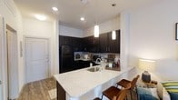 This single-floor space showcases a well-appointed kitchen designed for both functionality and style. The kitchen is anchored by a spacious peninsula topped with light granite countertops featuring subtle speckled detailing, offering ample prep space and seating for multiple bar-height chairs — ideal for casual dining or entertaining. An undermount sink is integrated seamlessly into the peninsula.

Dark espresso-toned cabinetry lines the kitchen walls, providing generous storage and creating a striking visual contrast against the white and neutral-toned walls and light tile backsplash. Black appliances, including a full-size refrigerator, microwave, and range, complement the dark cabinetry for a cohesive aesthetic.

Overhead, two elegant cylindrical pendant lights hang above the peninsula, while recessed lighting throughout ensures the space is well-lit. The flooring transitions from wood-look planks in the main area to tile near the entryway, adding visual interest and practicality. A white entry door is visible to the side, reinforcing the clean, contemporary palette of the space. Overall, the kitchen exudes a modern, polished ambiance.