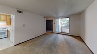 This single-floor residential unit offers a straightforward and functional layout designed for comfortable everyday living. The living area is generously sized, featuring neutral-toned carpet flooring, crisp white walls, and a sliding glass door that fills the room with natural light and provides a view of the outdoors. A pendant light fixture marks the transition toward the dining area, adding a subtle decorative touch. The kitchen is well-equipped with warm-toned wooden cabinetry extending to the ceiling for ample storage, white tile flooring, a gas range with an overhead vent hood, a refrigerator, and a stainless steel sink. Counter space is practical and efficiently laid out. The bedrooms are carpeted and bright, with one featuring a large window that overlooks a tree-lined outdoor area, and dark-stained interior doors throughout add a consistent design element. The unit includes two bathrooms: a full bath with beige square tile surrounding the tub-shower combination, tile flooring, a vanity with cabinetry, and a half bath with tile flooring and accessibility grab bars installed near the toilet. Overall, the space presents a clean, move-in ready environment with a cohesive neutral palette and practical finishes throughout.