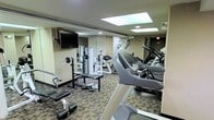 This fitness center is a compact yet fully functional exercise facility designed to accommodate a wide range of workout routines. The space is outfitted with a selection of cardio equipment, including treadmills and other aerobic machines positioned along one side of the room, providing users with ample options for cardiovascular training. On the opposite side, a free weights area features a bench press setup, weight plates, and a multi-function strength training machine, catering to those focused on resistance and strength workouts. A wall-mounted flat-screen television is positioned for easy viewing from most areas of the room, enhancing the workout experience. The floor is covered in a durable, patterned carpet suited for a fitness environment. Overhead, recessed lighting and skylight panels provide bright, even illumination throughout the space. Full-length mirrors along the walls help to visually expand the room and allow users to monitor their form during exercise. An emergency exit is clearly marked at the rear of the space, ensuring safety compliance. Overall, this fitness center is thoughtfully arranged to maximize utility within its footprint, making it a practical and well-appointed facility for daily exercise.