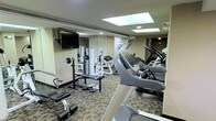 This fitness center is a compact yet fully functional exercise facility designed to accommodate a wide range of workout routines. The space is outfitted with a selection of cardio equipment, including treadmills and other aerobic machines positioned along one side of the room, providing users with ample options for cardiovascular training. On the opposite side, a free weights area features a bench press setup, weight plates, and a multi-function strength training machine, catering to those focused on resistance and strength workouts. A wall-mounted flat-screen television is positioned for easy viewing from most areas of the room, enhancing the workout experience. The floor is covered in a durable, patterned carpet suited for a fitness environment. Overhead, recessed lighting and skylight panels provide bright, even illumination throughout the space. Full-length mirrors along the walls help to visually expand the room and allow users to monitor their form during exercise. An emergency exit is clearly marked at the rear of the space, ensuring safety compliance. Overall, this fitness center is thoughtfully arranged to maximize utility within its footprint, making it a practical and well-appointed facility for daily exercise.