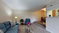 This single-floor residence showcases a thoughtfully designed layout with a distinctive split-level interior. The living room sits at a slightly lower elevation, creating a sunken effect that adds architectural character and a sense of intimacy to the space. Soft, neutral-toned carpeting flows throughout, lending warmth underfoot and tying the rooms together cohesively. The living area is generously proportioned, with smooth, light-colored walls and a flat ceiling that contribute to a clean, understated aesthetic. Ample room accommodates comfortable seating arrangements, and a glass-top coffee table adds a modern touch to the relaxed setting. A few steps lead up to the raised dining area, which is distinguished by a ceiling fan overhead and wood-toned flooring, creating a subtle yet effective visual separation between the two spaces. This elevated dining zone offers a natural gathering area, complete with a dining table and chairs. The neutral color palette maintained throughout the home provides a versatile backdrop suitable for a variety of décor styles. Overall, the space balances practicality with a warm, welcoming ambiance, making it well-suited for comfortable everyday living.