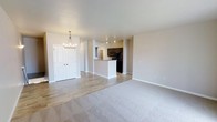 This single-floor space offers a thoughtfully designed open-concept layout that connects the living area and kitchen in a seamless, functional arrangement. The living area is finished with plush neutral-toned carpet, providing a warm and comfortable foundation, while the kitchen and entryway transition to smooth hardwood-style flooring that adds a clean, modern contrast.

A central kitchen island with a light stone countertop serves as the focal point of the culinary space, complemented by dark cabinetry and black appliances, including a refrigerator. Track lighting overhead ensures the kitchen is well-illuminated and practical for everyday use.

The entryway is welcoming, featuring a stylish chandelier with warm-toned bulbs that adds an elegant touch to the transitional space. A set of double white-panel doors provides generous closet storage near the entry, adding to the overall functionality of the layout.

The walls throughout are finished in a soft, neutral gray-white palette that reflects light naturally and creates a bright, airy ambiance. Clean white baseboards and understated trim details contribute to the space's polished, contemporary aesthetic. The open floor plan and cohesive design make this a versatile and inviting environment well-suited for both everyday living and entertaining.