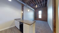 This single-story space showcases a striking industrial aesthetic rooted in its original architectural bones. The open-concept layout connects the kitchen and living areas seamlessly, creating a sense of expansive, uninterrupted space. Soaring ceilings with fully exposed wooden rafters and a prominent ventilation duct running overhead lend an authentic loft character. A large structural timber post anchors the interior, adding to the raw, warehouse-inspired atmosphere.

The kitchen features white shaker-style cabinetry, a peninsula with a dark countertop that provides additional prep and seating space, a stainless steel sink, and a dishwasher. Warm-toned pendant lights hang from the ceiling, casting focused illumination over the kitchen area and contributing to an inviting ambiance.

The living area transitions to plush carpet flooring, contrasting with the hardwood-style flooring in the kitchen. An exposed brick wall runs along one side, reinforcing the industrial character, while a tall, narrow window allows natural light to filter in. The combination of original materials and modern updates gives this space a distinctive, urban edge.