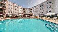 The courtyard and pool amenity at 40 Bennett Rd in Englewood, NJ, offers a thoughtfully designed outdoor retreat enclosed by a multi-story residential building. At its centerpiece is a generously sized swimming pool with crystal-clear water, surrounded by a spacious sun deck lined with white lounge chairs and shade umbrellas, providing ample space for relaxation. A covered pergola structure along the poolside offers a shaded seating area, perfect for enjoying the outdoors on warmer days. Adjacent to the pool, a herringbone-patterned brick paver courtyard hosts multiple modern outdoor dining tables and chairs beneath sleek overhead pergolas, creating an inviting al fresco gathering space. A built-in outdoor kitchen station, clad in natural stone and equipped with a stainless steel grill, adds functional appeal for outdoor entertaining. Manicured landscaping, including ornamental grasses, flowering plants, and neatly trimmed greenery, weaves throughout the courtyard, softening the hardscape and adding vibrant color. Decorative planters with lush foliage provide additional greenery near the covered seating zones. The overall ambiance is modern and resort-inspired, blending open-air leisure with well-appointed amenities in a cohesive, community-centered outdoor environment.