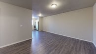 This single-floor unit offers a straightforward, functional layout designed for comfortable everyday living. The open living area is finished with durable dark wood-style vinyl flooring and neutral-toned walls, creating a clean and versatile space. A baseboard heater runs along the far wall, and ceiling-mounted lighting provides consistent illumination throughout. The living area flows directly into the kitchen, which is outfitted with white cabinetry, speckled light countertops, a double stainless steel sink, and a dishwasher, offering ample storage and prep space in a tidy, efficient arrangement. A stainless steel refrigerator and range are also visible from the main living area. The two bedrooms are carpeted in a neutral tone, each featuring a built-in closet with sliding or bifold doors, providing practical storage solutions. Walls throughout are painted in soft, light neutral tones that contribute to a calm and airy atmosphere. The bathroom is well-appointed with a single-sink vanity topped with a speckled countertop, a full tub-shower combination in white, dark wood-style vinyl flooring, and brushed nickel hardware accents. The overall design is clean, understated, and move-in ready, offering a practical and comfortable living environment.