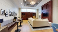 This refined single-floor space presents a thoughtfully designed lounge environment that balances elegance with functionality. The main lounge area is anchored by a coffered ceiling with a statement drum pendant light and recessed lighting throughout, casting a warm, welcoming ambiance over light hardwood flooring. A rich mahogany built-in shelving unit with an integrated clock serves as a focal point, flanked by open bookcase cabinetry and large windows that invite abundant natural light. The seating is varied and intentional — cream-toned sofas and accent chairs rest atop a geometric patterned area rug, while banquette-style bench seating with small pedestal tables lines one wall, offering more casual perches. A wall-mounted flat-screen television is framed within a bold mahogany surround, complemented by a mustard-yellow accent chair that adds a pop of color. An adjacent area features a full-sized grand piano alongside a long conference-style table with contemporary blue upholstered chairs, making the space equally suited to events and focused gatherings. Curated black-and-white photography and framed artwork adorn the walls, lending a cultured, gallery-like quality. Built-in storage and display shelving provide practical touches throughout, completing a space that is polished, versatile, and visually cohesive.