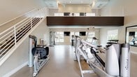 This well-appointed two-story fitness center offers a comprehensive workout environment designed to accommodate a wide range of exercise needs. The main floor features soaring ceilings and an open layout filled with professional-grade Hoist strength training machines, including cable systems, leg press, and multi-functional units. A dedicated free weights area includes a dumbbell rack, adjustable benches, and a plyo box, while three large wall-mounted mirrors provide form-checking visibility throughout. The space is anchored by large windows that invite ample natural light and offer views of an outdoor pool area, creating an energizing atmosphere. A ceiling fan and recessed lighting further enhance comfort on the main level. A sleek white staircase with a modern railing system leads to the upper mezzanine level, where carpeted flooring supports a row of Matrix treadmills, ellipticals, and stationary bikes. Mounted flat-screen televisions face the cardio equipment on both floors, and additional ceiling fans maintain airflow throughout. The upper level overlooks the main floor below through an open railing, reinforcing the facility's spacious, double-height design. Clean, neutral tones and polished tile flooring on the ground level contribute to the space's contemporary aesthetic.