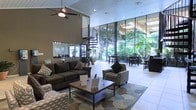 This single-story space makes a strong architectural statement through its dramatic floor-to-ceiling glass wall system, which spans the full width of the main living area and floods the interior with natural light. The towering ceilings amplify the sense of openness, and a ceiling fan is mounted overhead to complement the airy atmosphere. A striking spiral staircase serves as a sculptural focal point within the space, adding visual interest while connecting levels. The main living area is furnished with dark upholstered sofas and patterned accent chairs arranged around a low-profile wood and metal coffee table, all anchored by a large area rug with a soft, organic pattern. Large-format tile flooring extends throughout, contributing to the clean, contemporary aesthetic. Through the expansive glass wall, a covered outdoor area is visible, lined with tropical greenery and furnished with bar-height seating, suggesting a seamless indoor-outdoor connection. A mirrored or reflective wall element near the back of the space enhances depth and light distribution. Overall, the space balances modern design with a warm, inviting ambiance suited for both relaxed gatherings and social entertaining.