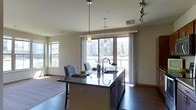 This single-floor space showcases a thoughtfully designed open-concept layout that seamlessly connects the kitchen and living areas. The kitchen serves as the centerpiece, anchored by a substantial island topped with dark granite countertops and an undermount sink, offering both functionality and visual appeal. Sleek pendant lights hang above the island, complementing a track lighting system that adds a modern, versatile touch to the space. The cabinetry and dark hardwood flooring throughout contribute to a sophisticated, contemporary aesthetic. Large windows and a sliding glass door flood the interior with abundant natural light while framing views of the surrounding neighborhood. The neutral color palette, featuring soft white walls and warm wood tones, creates a calm and inviting ambiance. Stylish upholstered bar stools along the island provide casual seating, making the kitchen a natural gathering spot. The open flow between the kitchen and adjacent living area enhances the sense of spaciousness, making this a well-appointed and practical space suited for both everyday living and entertaining.